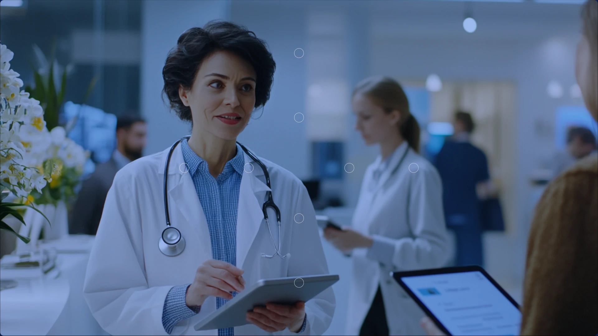 A female doctor with a stethoscope around her neck holding a tablet and speaking to a patient in a hospital setting.