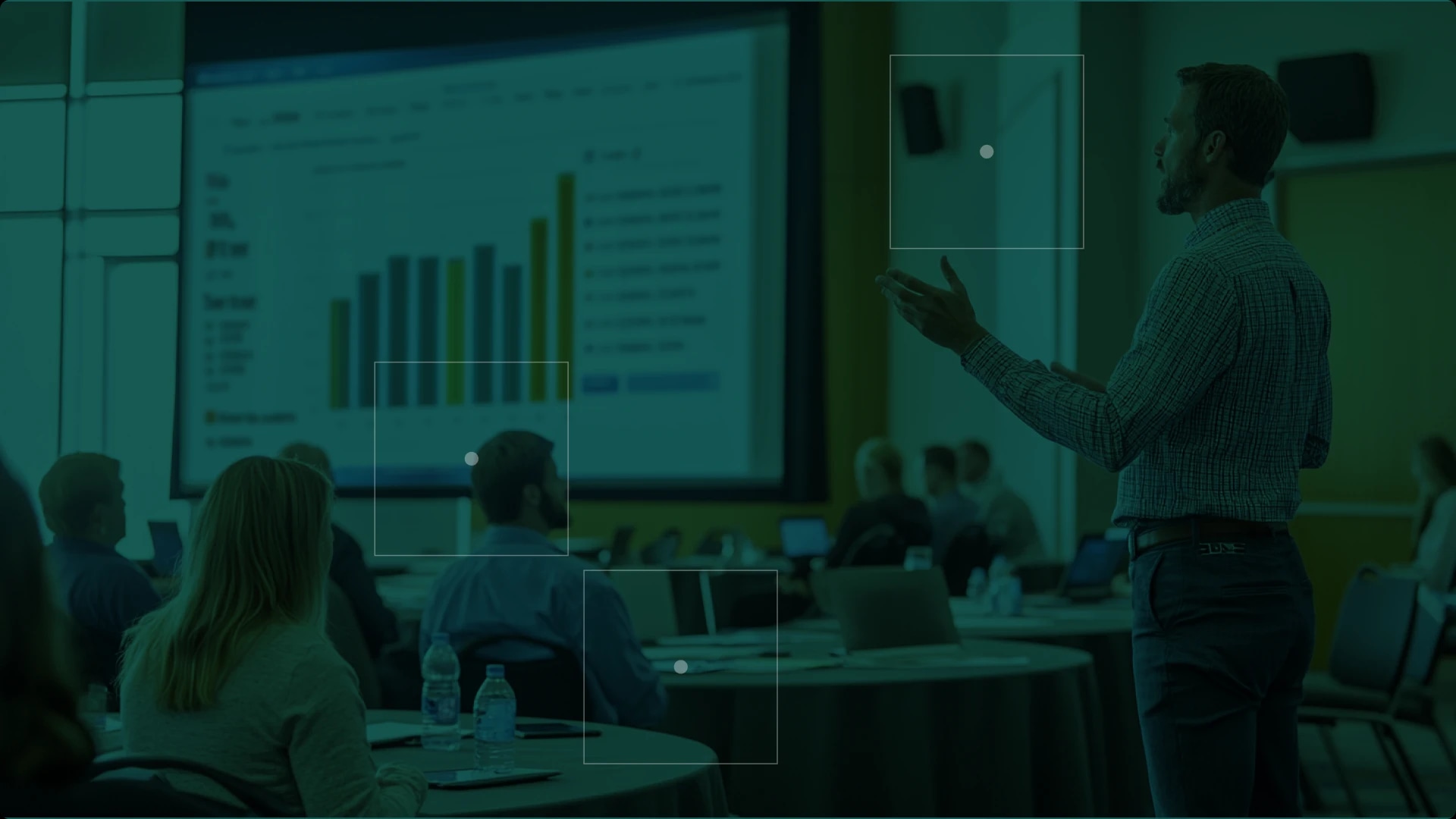A man giving a presentation to an audience in a conference room with a large screen displaying a bar chart.