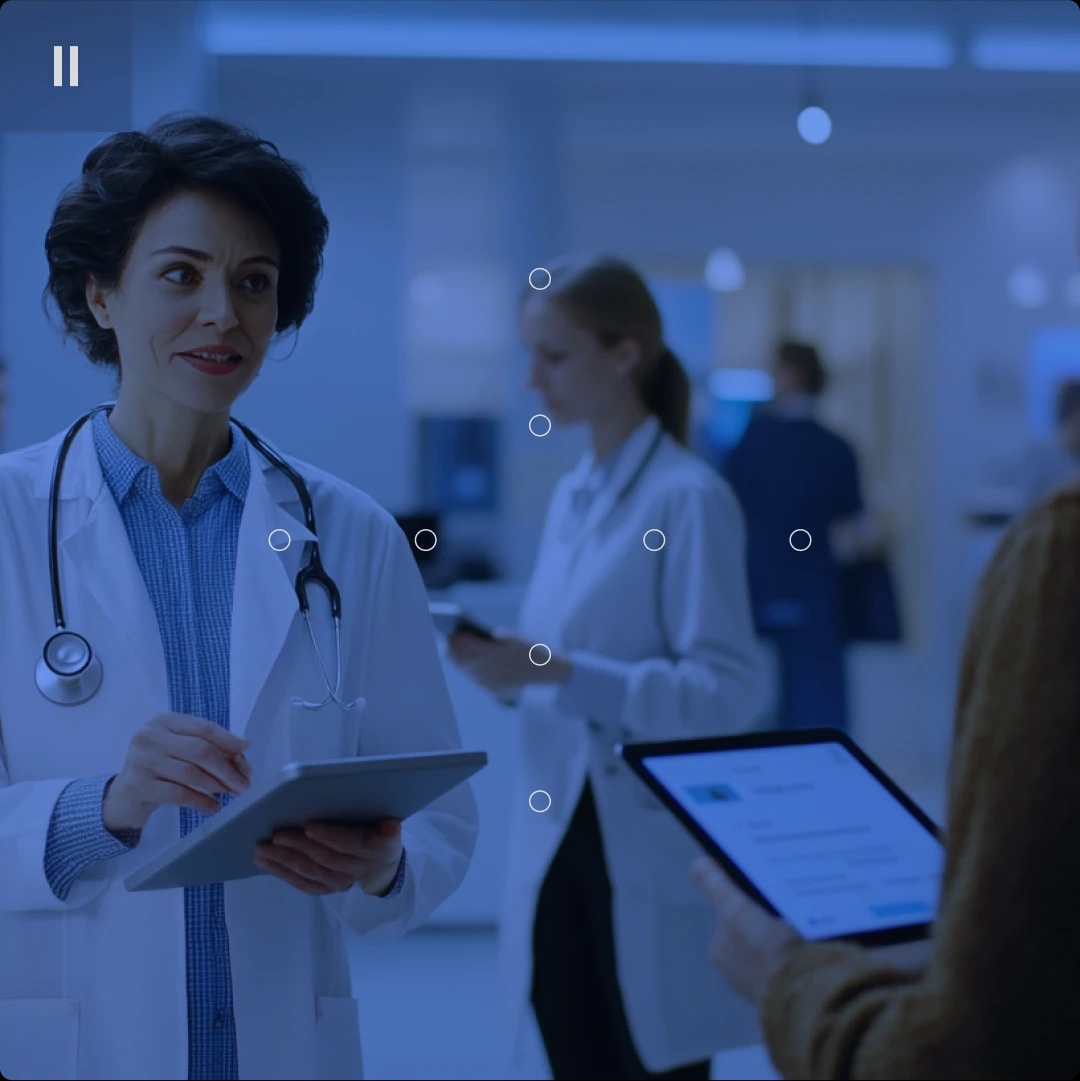 A female doctor with a stethoscope around her neck holding a tablet, talking to a person holding a tablet in a hospital setting.